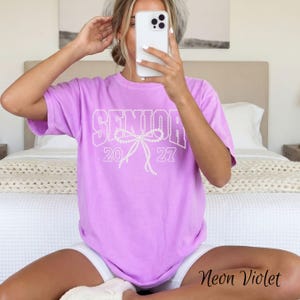 Puede incluir: Una camiseta de manga corta color violeta ne&oacute;n con el texto blanco SENIOR 2027 y un gr&aacute;fico de lazo central. El dise&ntilde;o est&aacute; compuesto por peque&ntilde;os puntos blancos, creando un efecto de contorno punteado en el pecho de la prenda.