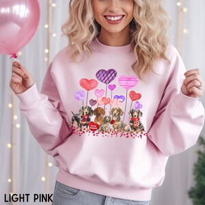 May include: Pale pink sweatshirt with a Valentine's Day theme. The design features dachshund dogs, heart-shaped balloons, and the word "LOVE". The phrase "Free Kisses" is also included. The sweatshirt is made of a soft, comfortable material.