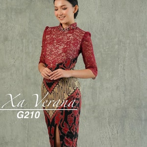 Op de afbeelding: Een rood-zwarte jurk met een kantbovenstuk en een rok met batikprint. De jurk heeft een hoge halslijn en een split in de rok. De jurk is gelabeld met "Xa Verana G210".