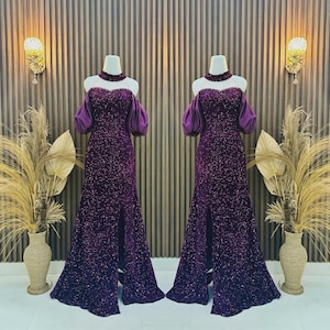 Puede incluir: Dos vestidos de noche largos en maniquíes. Los vestidos son de color púrpura intenso, cubiertos de lentejuelas, con cuello alto y mangas descubiertas. Cada vestido tiene una abertura en el muslo. El fondo presenta apliques de pared decorativos y jarrones.