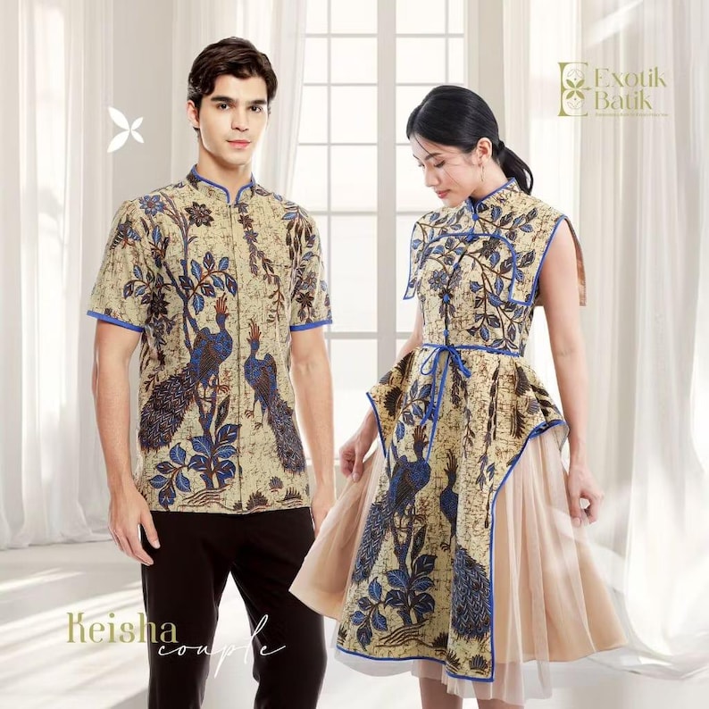 May include: Matching beige and blue batik outfits. The man wears a short-sleeved shirt with a peacock design, paired with black pants. The woman wears a dress with a similar peacock pattern and a beige tulle skirt. The outfits are from Exotik Batik.