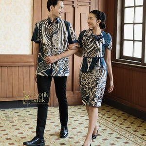 Puede incluir: Una pareja con conjuntos de batik a juego. El hombre lleva una camisa de manga corta y pantalones negros. La mujer lleva un top y una falda lápiz a juego. Los conjuntos presentan un estampado floral azul marino, blanco y beige. El logotipo "Exotik Batik" es visible.