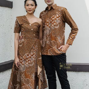 Puede incluir: Una mujer con un vestido marrón de un solo hombro y un hombre con una camisa con botones estampada marrón y pantalones negros. La ropa presenta un patrón detallado y repetitivo en tonos marrones y beige. El vestido tiene un diseño de falda cruzada.