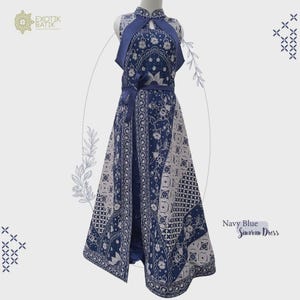 Op de afbeelding: Marineblauwe Sinaran-jurk met een uniek batikpatroon. De jurk heeft een halterhalslijn, een bijpassende riem en een split aan de voorkant. Het is versierd met bloemen en geometrische ontwerpen, en de tekst "Exotik Batik" is zichtbaar.