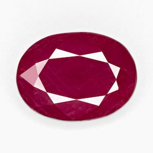 Puede incluir: Gema de rubí de forma ovalada con talla facetada. La piedra de color rojo intenso tiene facetas geométricas blancas que reflejan la luz. La gema está sobre un fondo blanco.