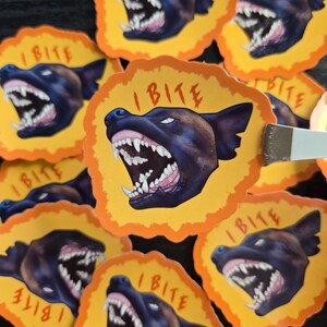 Peut inclure: Une collection d'autocollants découpés représentant une tête de chien noir grognant avec les dents découvertes et le texte I BITE en lettres rouges. Chaque autocollant a un centre jaune et une bordure orange irrégulière.