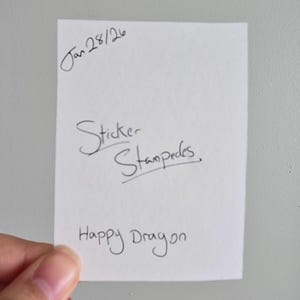 Op de afbeelding: Een wit rechthoekig papier met handgeschreven tekst. De tekst bevat "Jan 28/26", "Sticker Stampedes" en "Happy Dragon". Het papier wordt door een hand tegen een effen witte achtergrond gehouden.