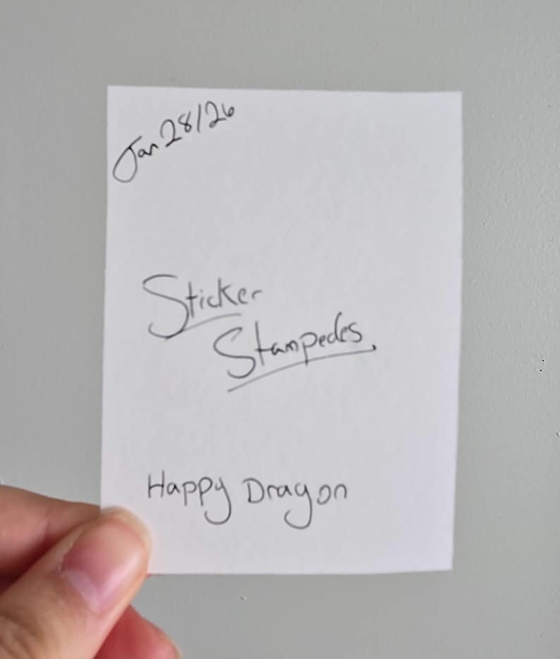 Op de afbeelding: Een wit rechthoekig papier met handgeschreven tekst. De tekst bevat "Jan 28/26", "Sticker Stampedes" en "Happy Dragon". Het papier wordt door een hand tegen een effen witte achtergrond gehouden.