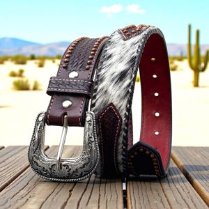 May include: A brown leather belt with a silver buckle and a section of black and white cowhide. The belt has decorative studs and a red interior. The buckle is ornate with floral designs. The belt is displayed on a wooden surface.