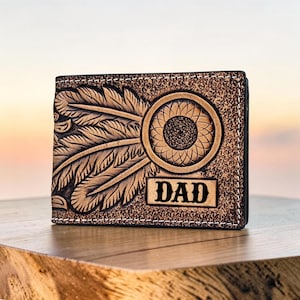Könnte beinhalten: Eine braune Ledergeldbörse mit Sonnenblumen- und Federmuster. Das Wort "DAD" ist auf der Vorderseite eingeprägt. Die Geldbörse liegt auf einer Holzoberfläche.