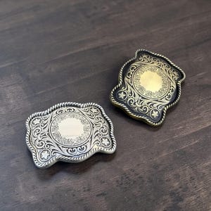 May include: Two decorative belt buckles with ornate scrollwork and star accents. One buckle is silver-toned, the other is gold-toned with a black border. Both have a rope-like edge and a central circular design.