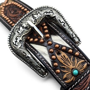May include: Close-up of a Western-style belt. The belt features a silver-toned buckle with intricate scrollwork. The black leather strap has a white and brown hair-on hide design, copper-toned studs, and a turquoise stone accent.
