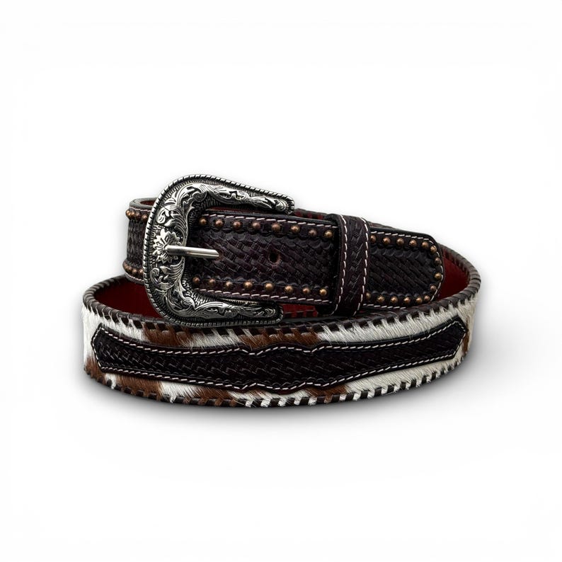 May include: A brown leather belt with a silver buckle. The belt features a woven pattern and is accented with small metal studs. The lower portion of the belt has a cowhide design with brown, white, and black patches.