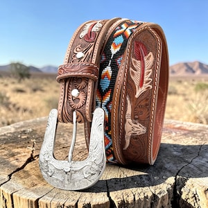 May include: A brown leather belt with a silver buckle featuring a steer head design. The belt has intricate carved details, including feathers and floral patterns. A second belt with colorful beadwork is also visible.