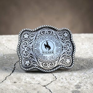 May include: Silver-toned belt buckle with intricate scrollwork and a central circular design. The design features a silhouette of a cowboy on a horse, with the name "WALKER" below. The buckle has a rope-like border and star accents.