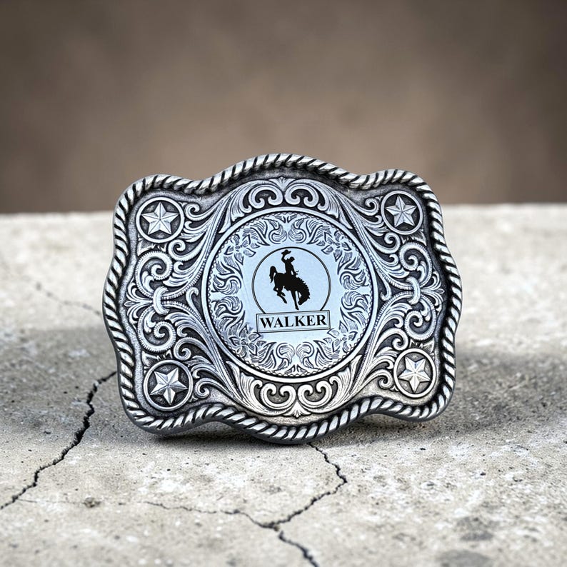 May include: Silver-toned belt buckle with intricate scrollwork and a central circular design. The design features a silhouette of a cowboy on a horse, with the name "WALKER" below. The buckle has a rope-like border and star accents.