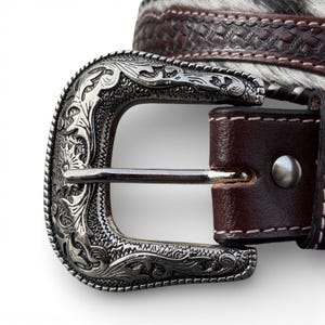May include: Close-up of a western-style belt with a silver-colored buckle. The buckle has an ornate floral design. The belt is brown leather with a textured pattern and a section of black and white animal hide.