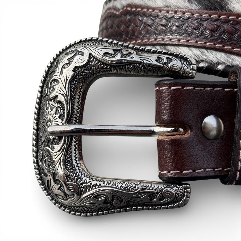 May include: Close-up of a western-style belt with a silver-colored buckle. The buckle has an ornate floral design. The belt is brown leather with a textured pattern and a section of black and white animal hide.