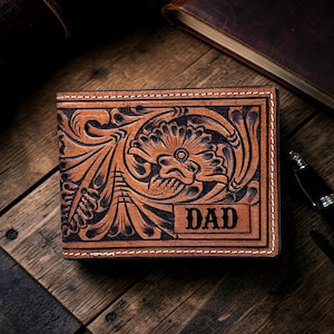 Könnte beinhalten: Braune Lederbrieftasche mit aufwendiger Blumen- und Totenkopf-Schnitzerei. Das Wort "DAD" ist auf einem rechteckigen Feld geprägt. Weiße Nähte. Die Brieftasche liegt auf Holz.