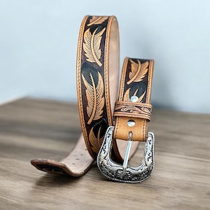 May include: A brown leather belt with a silver buckle. The belt is decorated with a pattern of black and brown feathers.
