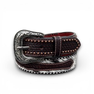 May include: A brown leather belt with a decorative silver buckle. The belt features a woven texture, copper-colored studs, and a section of black and white cowhide. The belt measures approximately 1.5 inches wide.