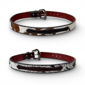 May include: Two leather belts with cowhide and dark brown accents. The belts have a silver buckle and a dark red interior. The cowhide has brown and white patches. The belts are displayed on a white background.