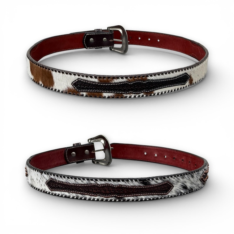 May include: Two leather belts with cowhide and dark brown accents. The belts have a silver buckle and a dark red interior. The cowhide has brown and white patches. The belts are displayed on a white background.