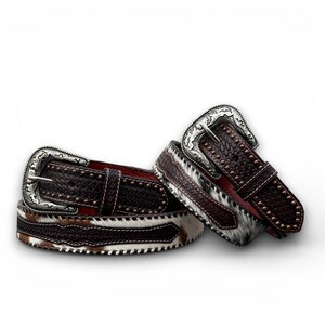 May include: Two brown leather belts with silver buckles. One belt features a woven pattern and small studs, while the other has a cowhide design with white and brown fur. Both belts have a red interior.