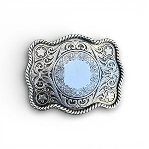 May include: Silver-colored rectangular belt buckle with intricate floral and scrollwork designs. The buckle features a central circular area and four star accents. A rope-like border frames the edges.