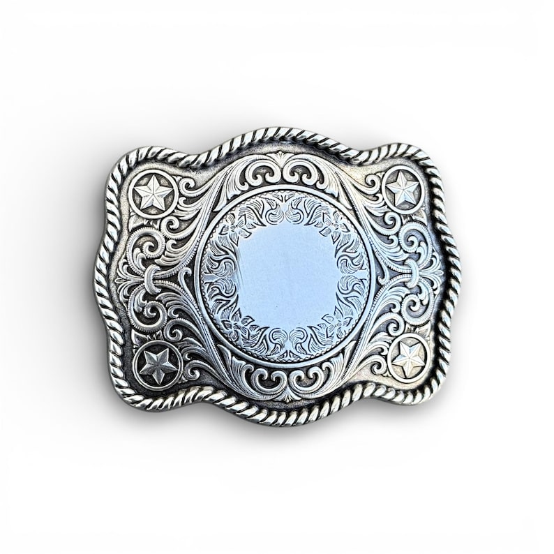 May include: Silver-colored rectangular belt buckle with intricate floral and scrollwork designs. The buckle features a central circular area and four star accents. A rope-like border frames the edges.