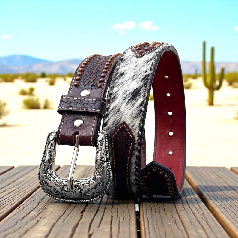 May include: A brown leather belt with a silver buckle and a section of black and white cowhide. The belt has decorative studs and a red interior. The buckle is ornate with floral designs. The belt is displayed on a wooden surface.