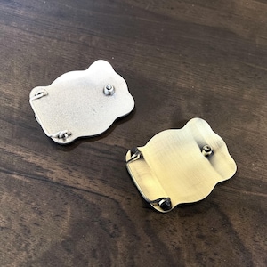 May include: Two metal belt buckles with a unique, organic shape. One is silver-toned, the other is gold-toned. Each buckle has a central post and small loops for attachment. The buckles are displayed on a dark wood surface.