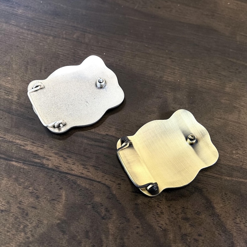May include: Two metal belt buckles with a unique, organic shape. One is silver-toned, the other is gold-toned. Each buckle has a central post and small loops for attachment. The buckles are displayed on a dark wood surface.