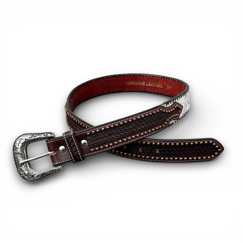 May include: Brown leather belt with a silver buckle. The belt features a woven texture, studded accents, and a red interior. The buckle has an ornate design. The belt is labeled "GENUINE LEATHER".