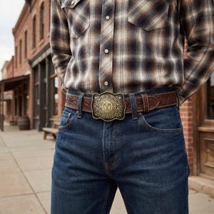May include: A brown leather belt with an ornate, gold-colored buckle. The belt is worn with blue jeans and a plaid shirt. The buckle features a central emblem surrounded by decorative scrollwork. The belt is a classic Western style.