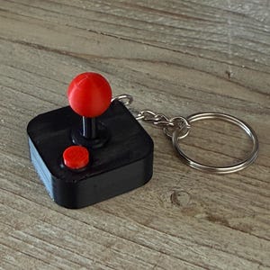 May include: A black square keyring shaped like a retro arcade joystick controller. The device features a prominent red spherical knob on a black shaft and a single circular red button on the top surface, attached to a silver metal split ring.