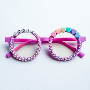 May include: Pink round glasses with rhinestones around the frames. The glasses have the name "ADDIE" spelled out in white beads on the top of the frame. The glasses also have a rainbow of small heart-shaped beads on the top of the frame.