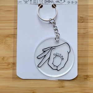May include: A clear acrylic keychain with a black outline of a hand making the "gotcha" gesture. The word "GOTCHA" is written inside the circle formed by the hand.
