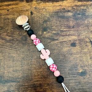 May include: A pink and black silicone baby teether with a wooden clip and a cactus charm. The teether has a checkered pattern and a pink and white cactus charm.