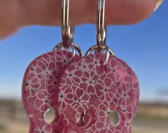 May include: Pink skull earrings with a floral design. The earrings have a clear, glossy finish and silver hoops. The skull-shaped pendants are decorated with white flowers and small black dots. The earrings are shown against a blue sky.
