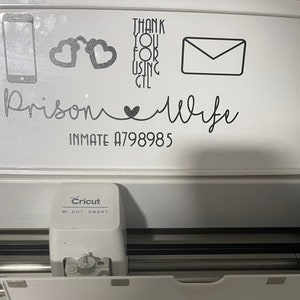 以下が含まれることがあります： 白い表面に、「Cricut をご利用いただきありがとうございます」と「Prison Wife Inmate A798985」という黒いテキストが書かれています。また、電話、手錠、封筒、ハートの画像もあります。表面は白い Cricut カッティングマシンの上にあります。