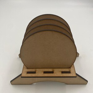 May include: A set of four unfinished wooden coasters with a wooden stand. The coasters are round and have a smooth surface. The stand has a simple design with four legs and a flat top.