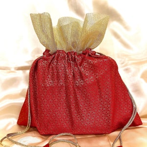 May include: A red drawstring bag with a gold mesh top and a gold cord. The bag is sitting on a cream-colored satin fabric.