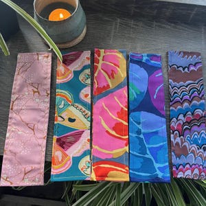May include: Five fabric bookmarks with colorful patterns. The first bookmark is pink with a tree and deer design. The second is teal with a butterfly design. The third is pink with a floral design. The fourth is blue with a leaf design. The fifth is brown with a wavy design.