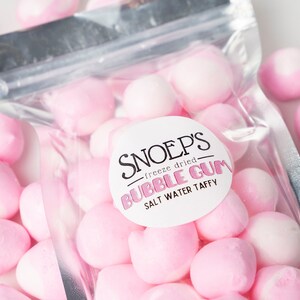May include: A silver resealable bag filled with pink freeze-dried bubble gum. The bag has a white label that reads "Snoep's - freeze dried - Bubble Gum - Salt Water Taffy".