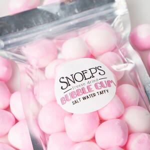 May include: A clear plastic bag filled with pink and white freeze-dried bubble gum. The bag has a label that reads "Snoep's - freeze dried - Bubble Gum - Salt Water Taffy".