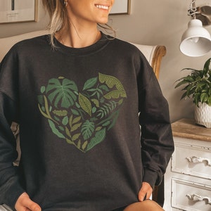 Puede incluir: Sudadera negra con un diseño de corazón de hojas verdes. El corazón está formado por varias hojas y plantas.