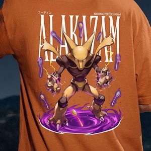 Pode incluir: Camiseta laranja com uma ilustração estilizada de Alakazam, um personagem Pokémon. O design inclui o nome "ALAKAZAM" em branco, com o texto "NATIONAL POKEDEX #0064" acima. O personagem é retratado com colheres e energia roxa.