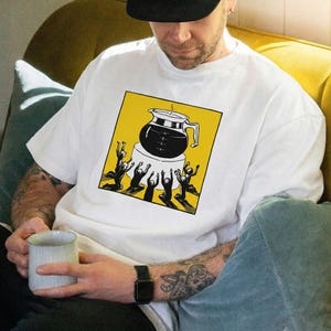Op de afbeelding: Wit T-shirt met een gele vierkante afbeelding met een zwart koffiepotontwerp en zwarte figuren met opgeheven armen. De persoon op de afbeelding draagt een zwarte pet en houdt een lichtblauwe mok vast.
