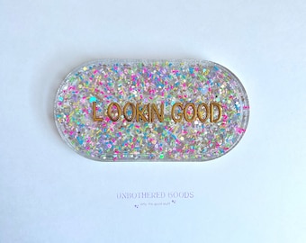 Puede incluir: Una bandeja ovalada transparente llena de purpurina colorida y el texto dorado "LOOKIN GOOD". La bandeja tiene un borde transparente y está sobre un fondo blanco. Las palabras "UNBOTHERED GOODS" están impresas en morado en la parte inferior.